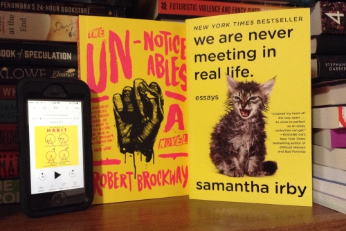 Image displays two books and an iPhone SE standing face out on a wooden bookshelf. There are several stacks of books in the background. The iPhone displays the cover of The Power of Habit on the Audible app (a yellow cover with four red hampster wheels and a stick man running in each one). The two books are The Unnoticeables and We Are Never Meeting in Real Life (a yellow cover with bold black writing and a kitten, who looks to be recovering from being in water, hissing).