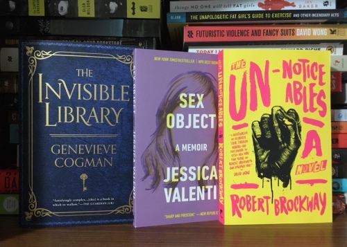 This image displays three books standing, covers facing out, on a wooden bookshelf. Behind these three books are stacks of more books. The three featured books are The Invisible Library (a blue cover with gold writing), Sex Object (a purple cover with bold white letters, featuring the head of a woman with long hair but no visible facial features), and The Unnoticeables (a yellow cover with bold pink writing, featuring a clenched fist).