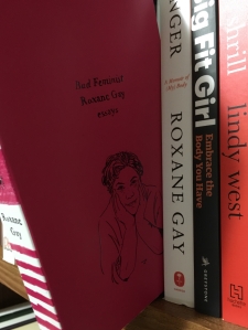 This image displays Bad Feminist, a hot pink book with a stylized drawing of Roxane Gay on the cover, leaning off the edge of a wooden bookshelf. Tucked properly on the shelf is also Gay's memoir, Hunger.