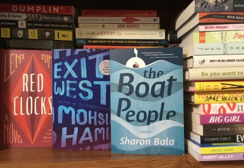 Image displays a wooden bookshelf containing the ever-growing To Be Read piles of books. In the foreground are three books with their covers facing out: Red Clocks (a red cover with overlapping red and purple diamond shapes behind white text), Exit West (a deep blue cover with light blue text; the letters have the uneven texture of something painted with a rough brush), and The Boat People (a predominantly light blue cover, depicting large waves with the title floating on them and a small red boat against a sunrise in the top left corner).