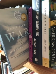Image displays a row of books on a wooden bookshelf. American War is pulled out and tilted slightly over the edge.